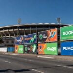 Painéis de apostas esportivas em avenida brasileira com estádio ao fundo